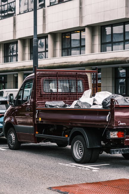 A small, dark red flatbed pickup truck parked on a city street in front of a modern, multi-story office building with large glass windows. The truck's cargo area is loaded with several large, white an