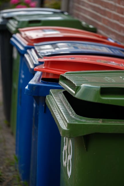 Close-up of stacked black plastic rubbish bags with shiny, crinkled surfaces, placed against a plain light-colored wall indoors or in a storage area. The bags are filled with waste and are tied at the